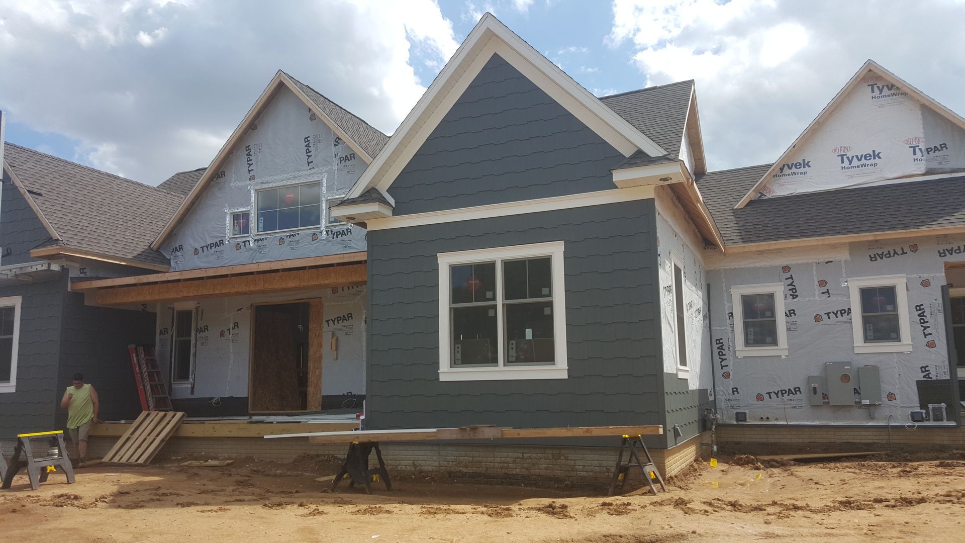Partially built suburban house with dark siding, white trim, and unfinished front yard under a cloudy sky