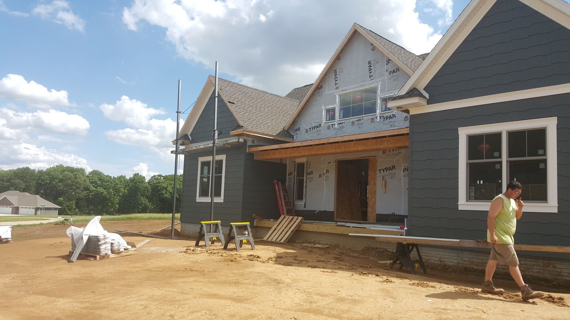 House under construction with gray siding, exposed roof beams, and a person standing on a dirt lot.