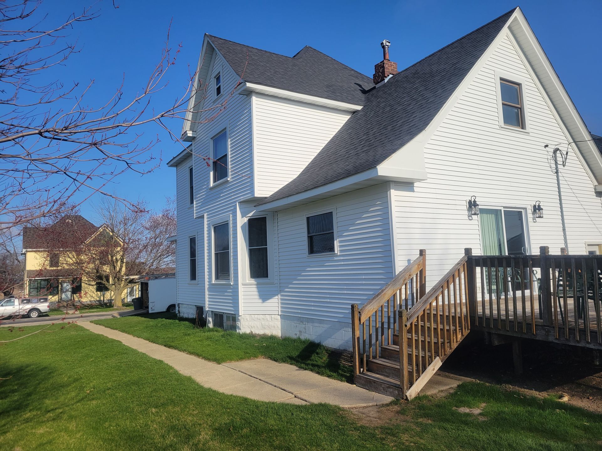 White two-story house with a small porch and wooden steps beside a sidewalk on a sunny day