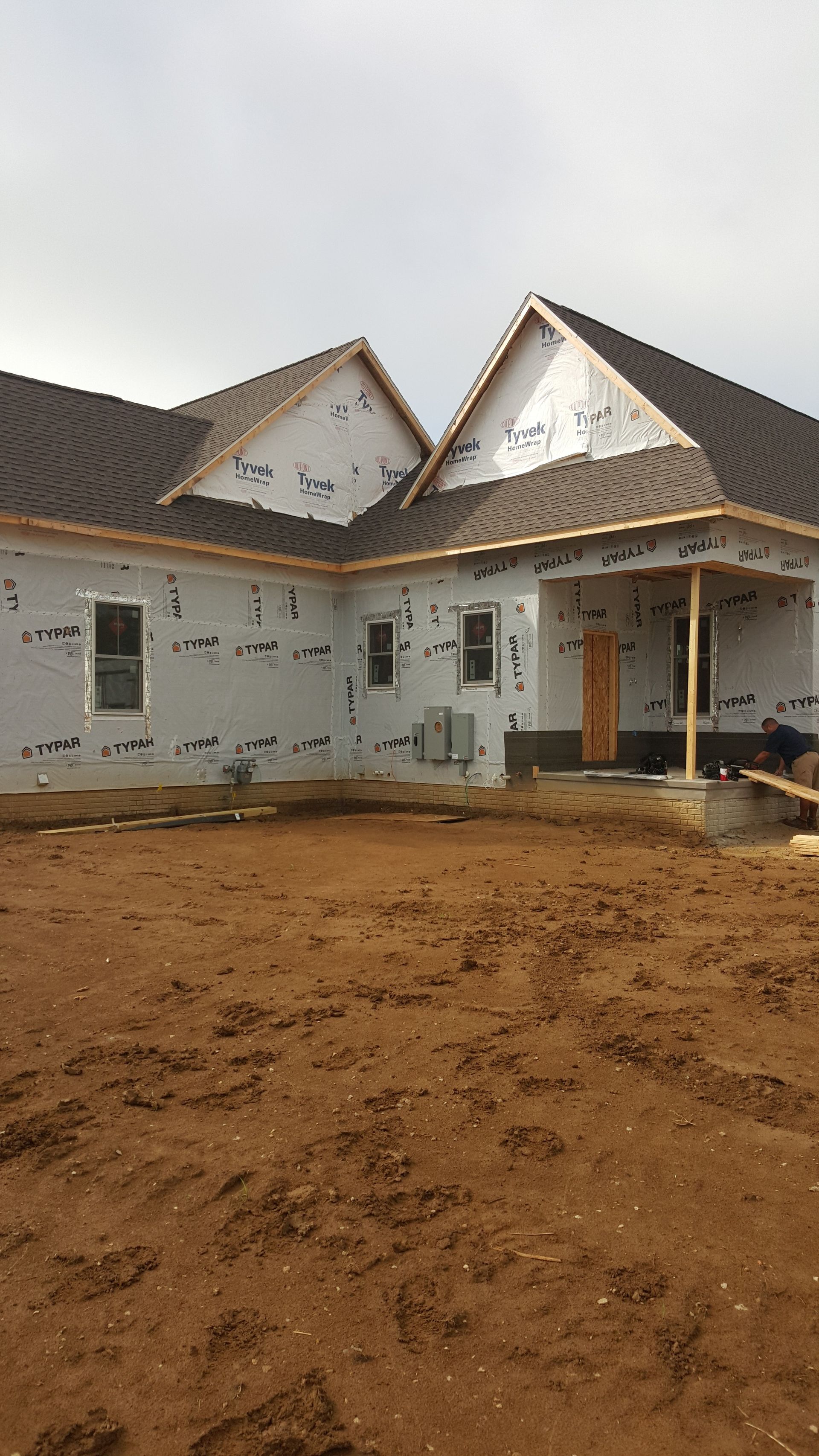 Partially built house with white walls and brown roof on a dirt lot under cloudy sky