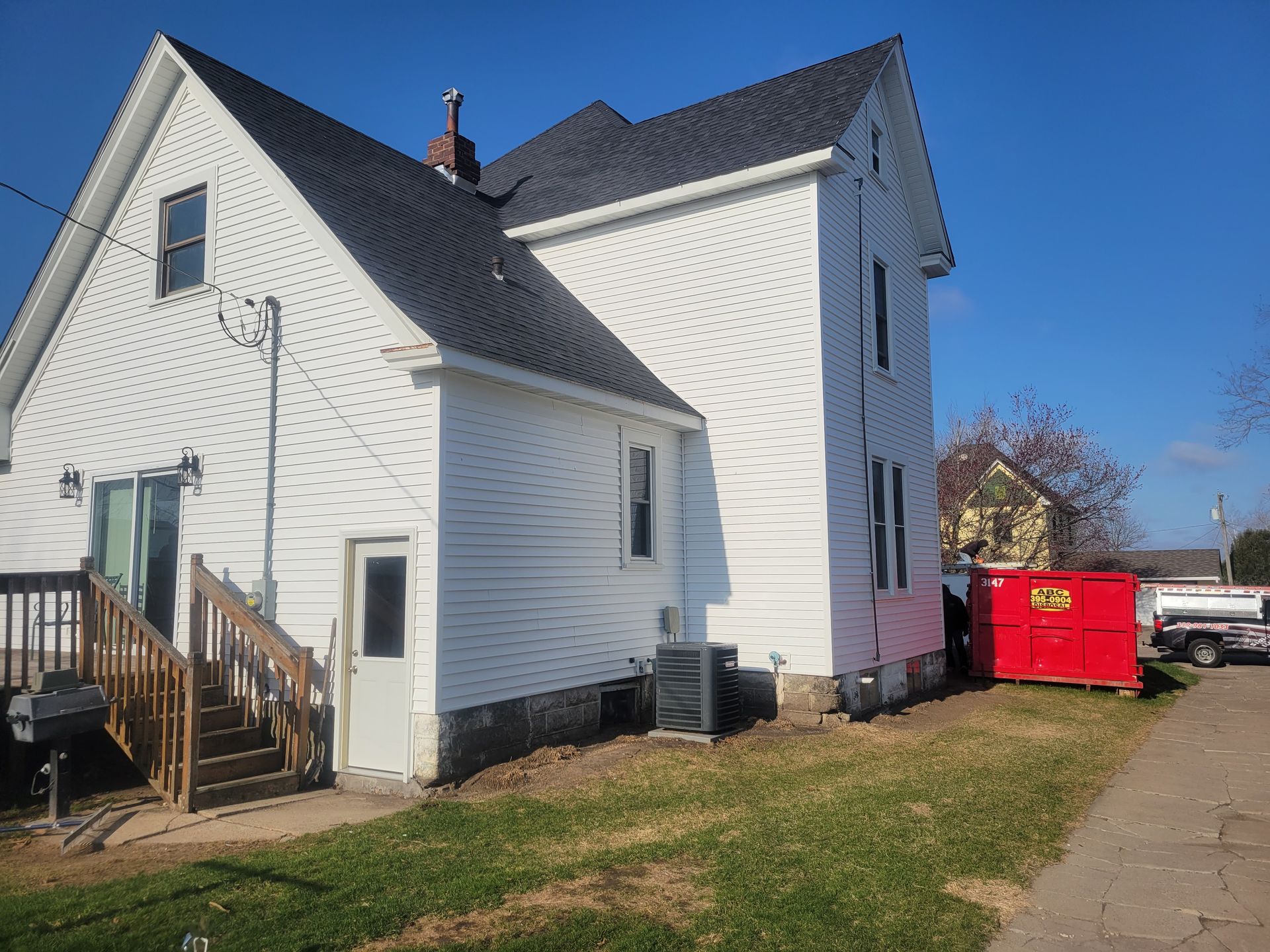White house with steep roof, wooden porch, and red dumpster beside a driveway on a sunny day