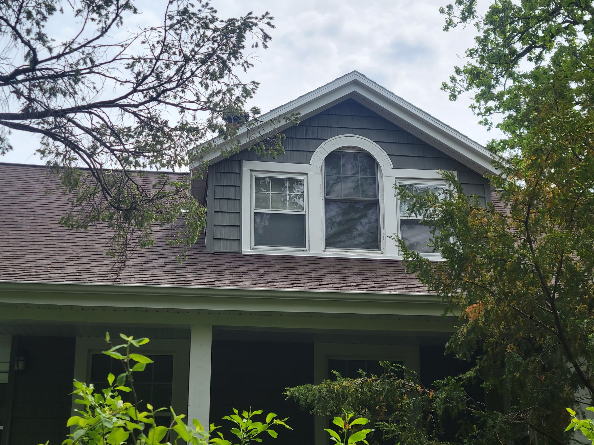 Front of a gray house with a dormer window and porch, framed by trees and branches