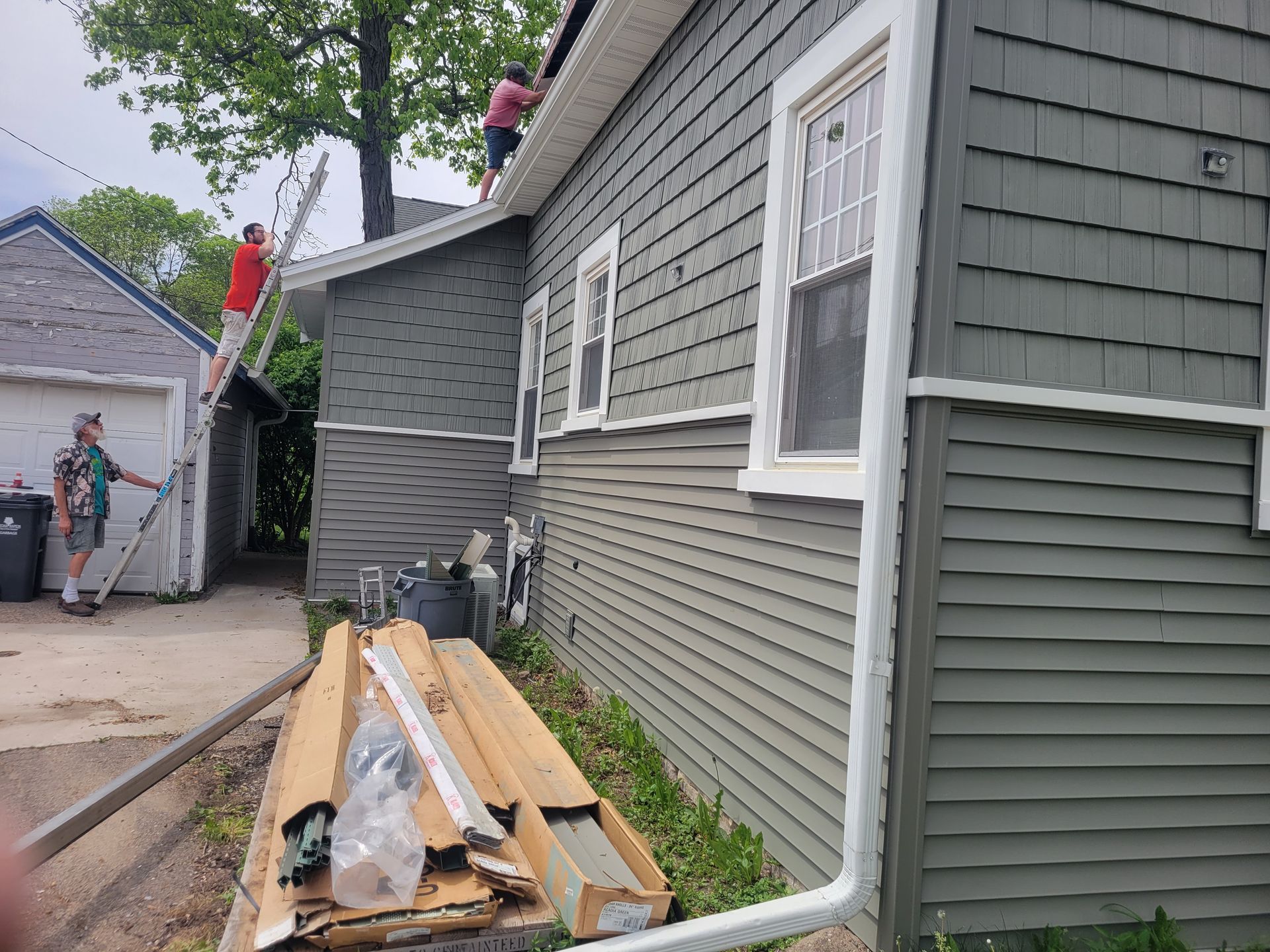Side of a gray house with stacked lumber and tools along a narrow driveway near a detached garage.