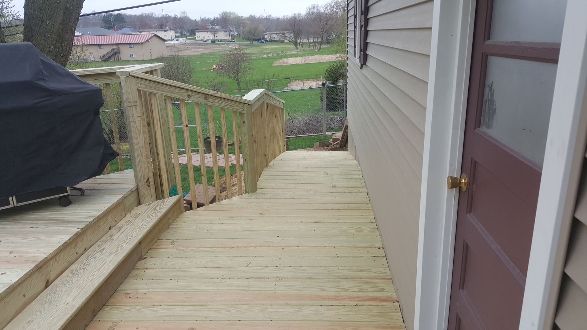Wooden deck beside a house with stairs, railing, and a grill overlooking a grassy backyard.