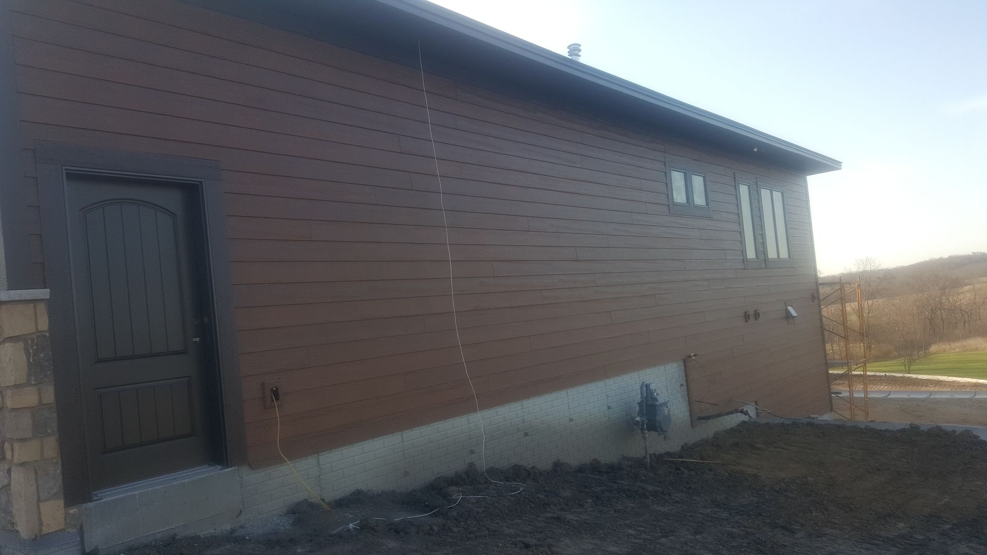Side view of a brown wooden building with a dark door and small windows, beside a gravel yard.