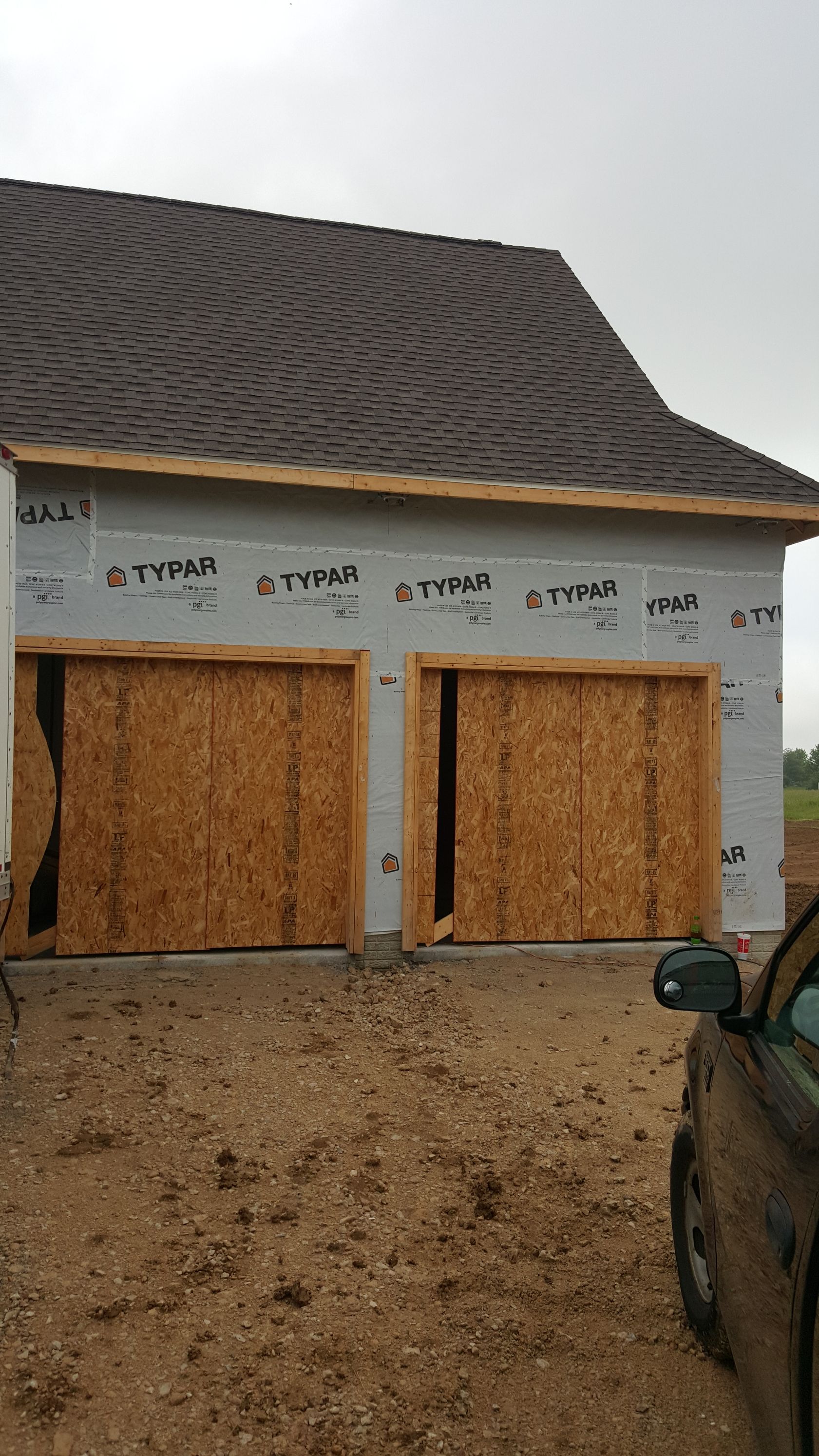Partially constructed house with boarded garage openings and a dark shingled roof under overcast sky