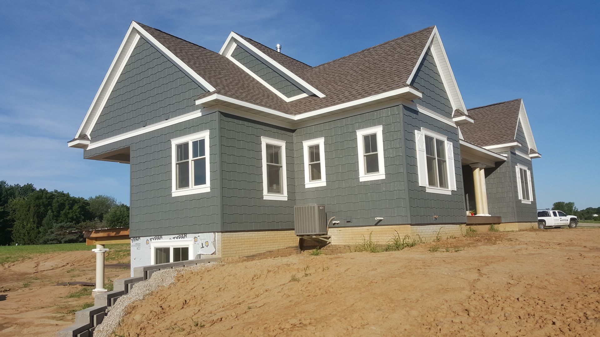 Gray house under construction with white trim and roof on a dirt lot under a clear blue sky