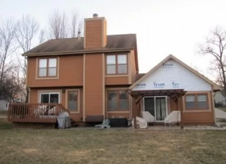 Two-story tan house with a brick chimney and a white-roofed porch on a grassy lot