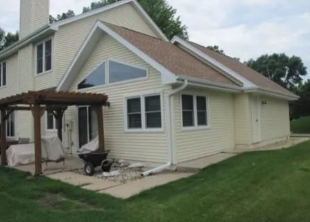 Beige house with a wooden pergola and stroller on a patio beside a grassy yard
