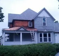 Two-story house with gray siding and a front porch; ladder leans against the roof under repair