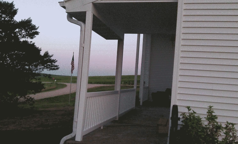 Porch of a white house overlooking a lawn and road at dusk, with an American flag in the distance.