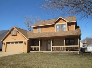 Suburban two-story tan house with attached garage and front porch under a clear blue sky