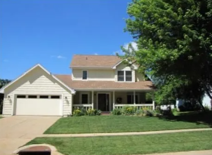 Suburban two-story house with beige siding, garage, front porch, and a green lawn under a blue sky