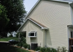 Suburban house with beige siding, small windows, and a sloped roof beside trees and a driveway