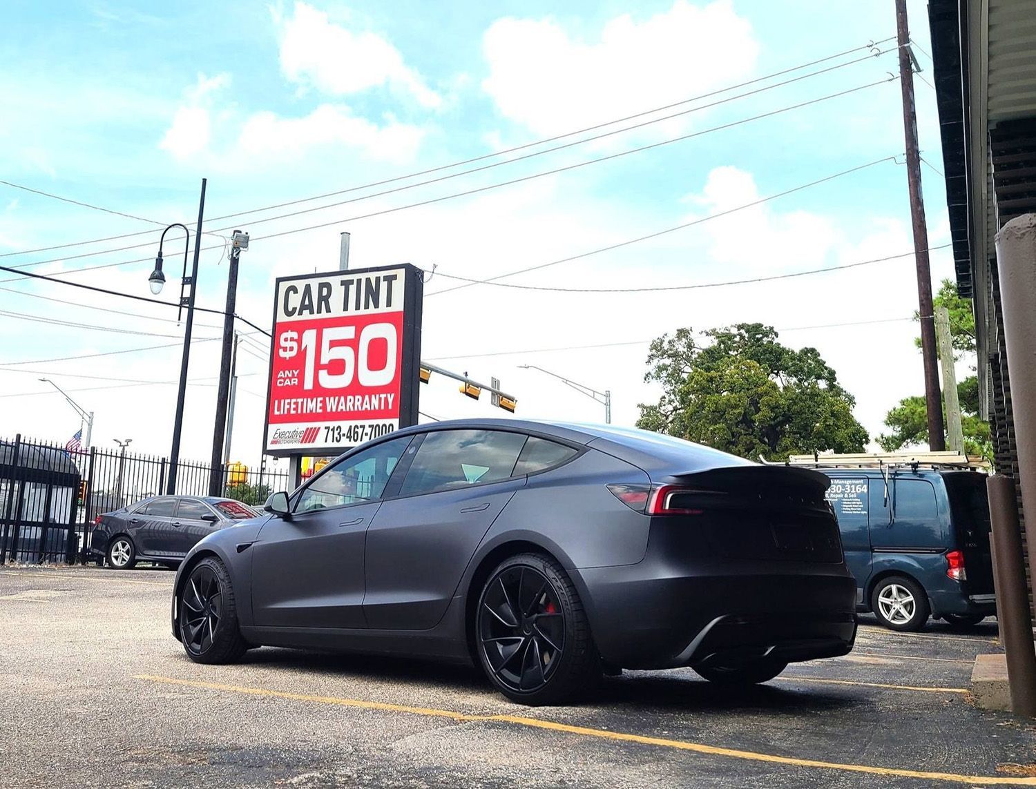 A tesla model 3 is parked in a parking lot in front of a car tint sign.