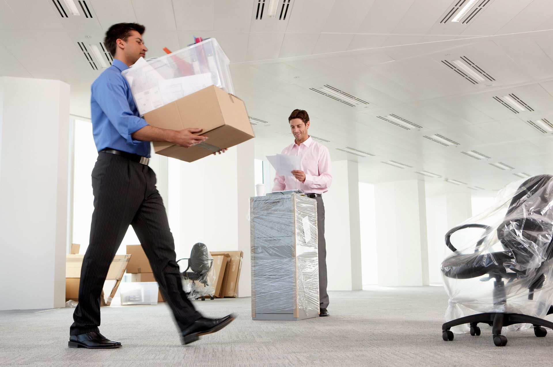 Two men moving boxes in a bright office, with a chair and wrapped furniture in the background.