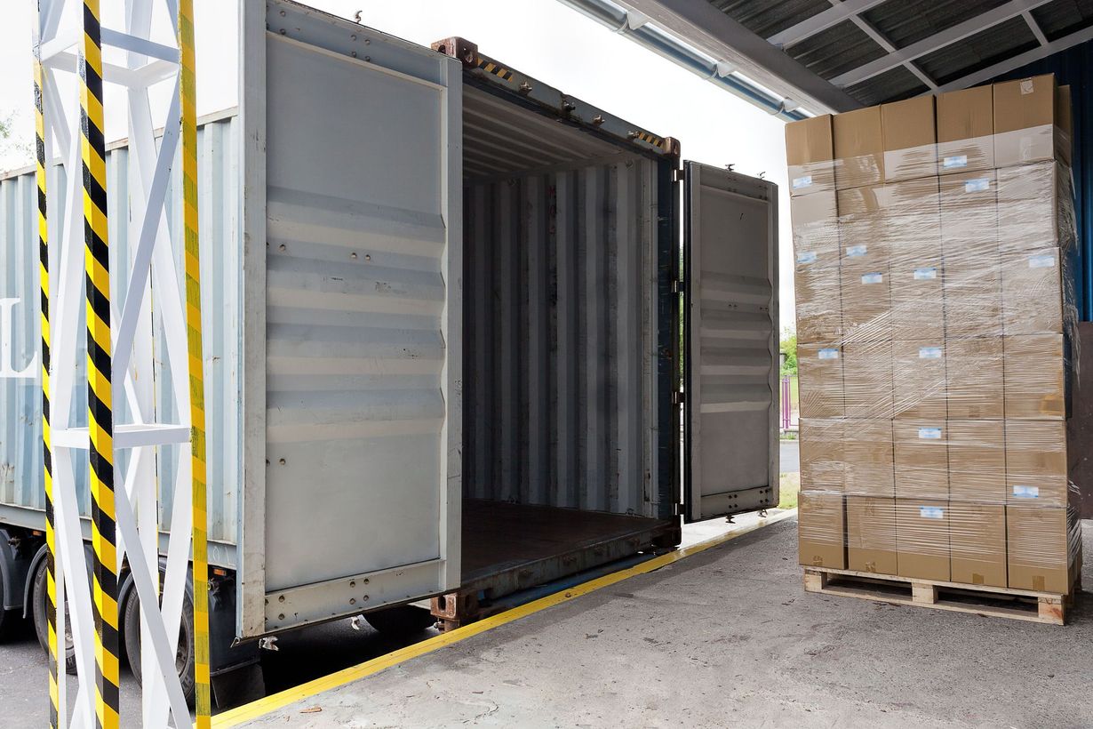Open shipping container beside stacked pallet of boxed goods in a warehouse loading area