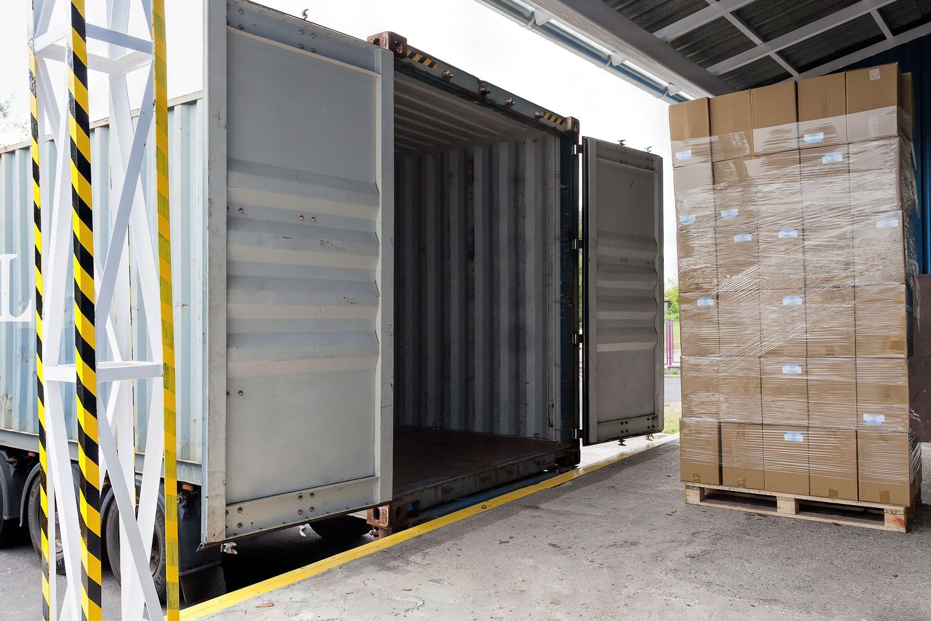 Open shipping container beside stacked pallet of boxed goods in a warehouse loading area
