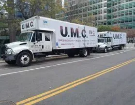 Two white U.M.C. moving trucks parked along a city street beside a glass office building.