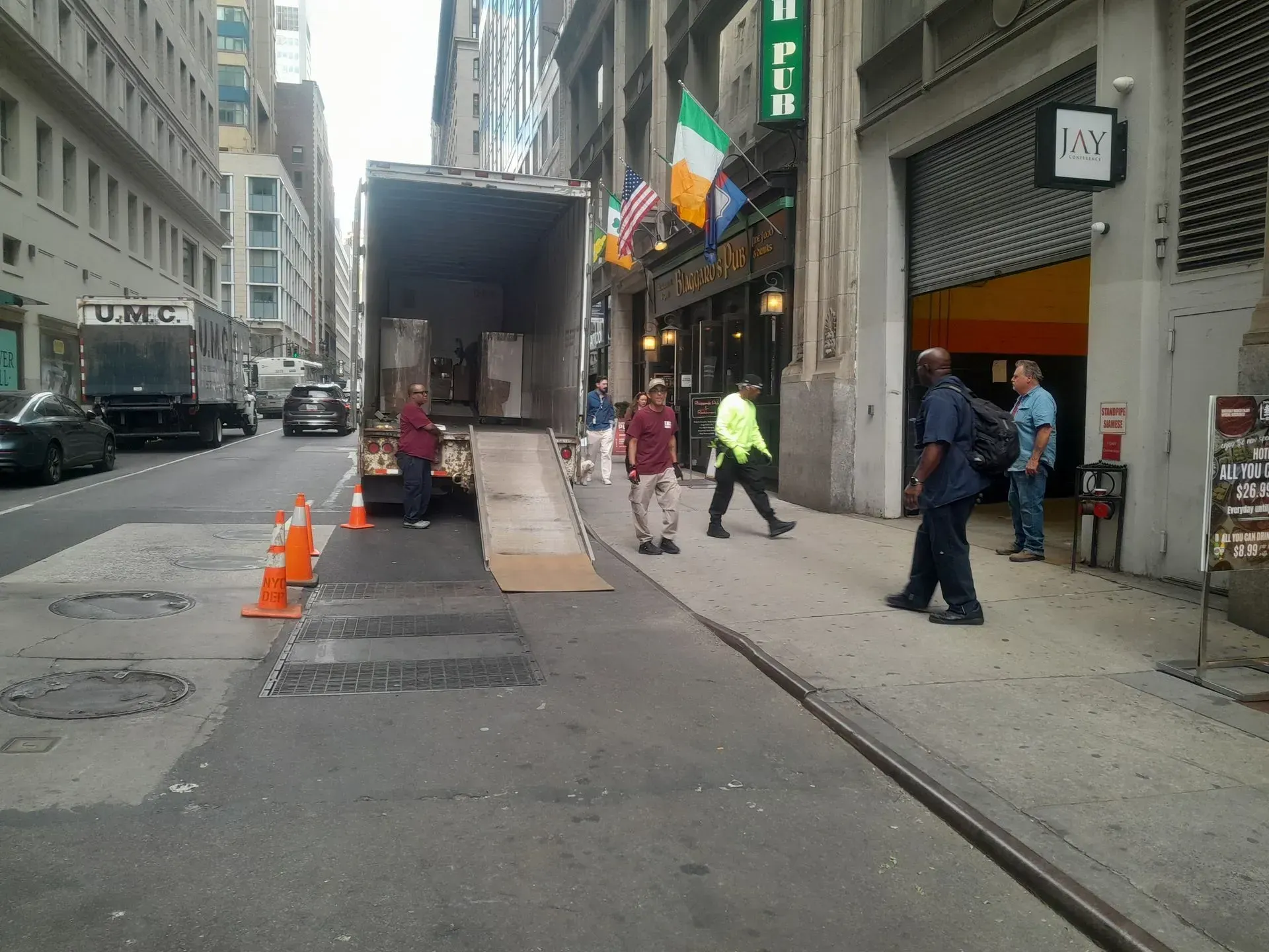 City sidewalk with workers, orange cones, and a truck unloading outside shops.