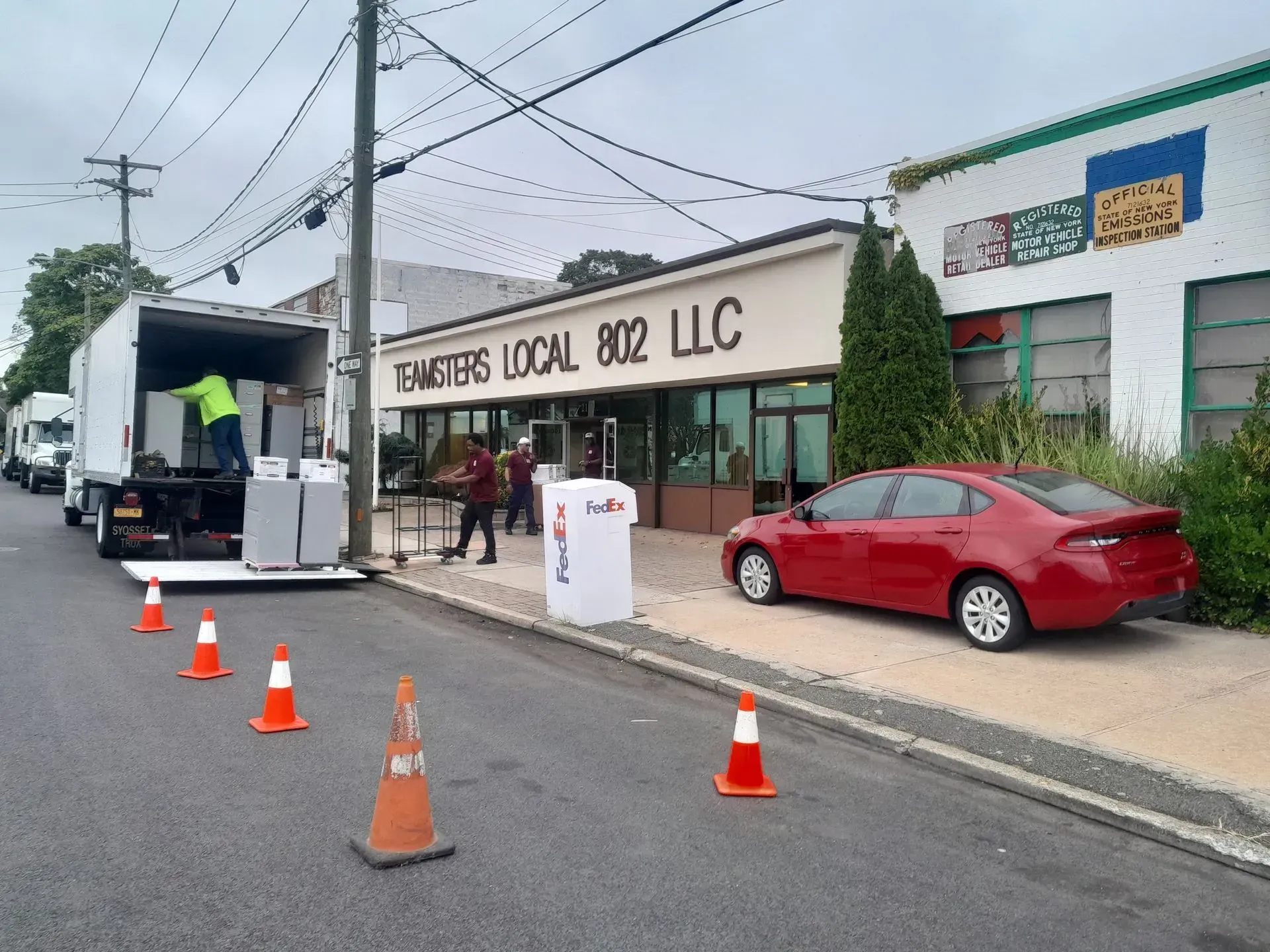 Workers unloading a truck outside Barnes Local 802 LLC, with cones and a red car parked nearby.