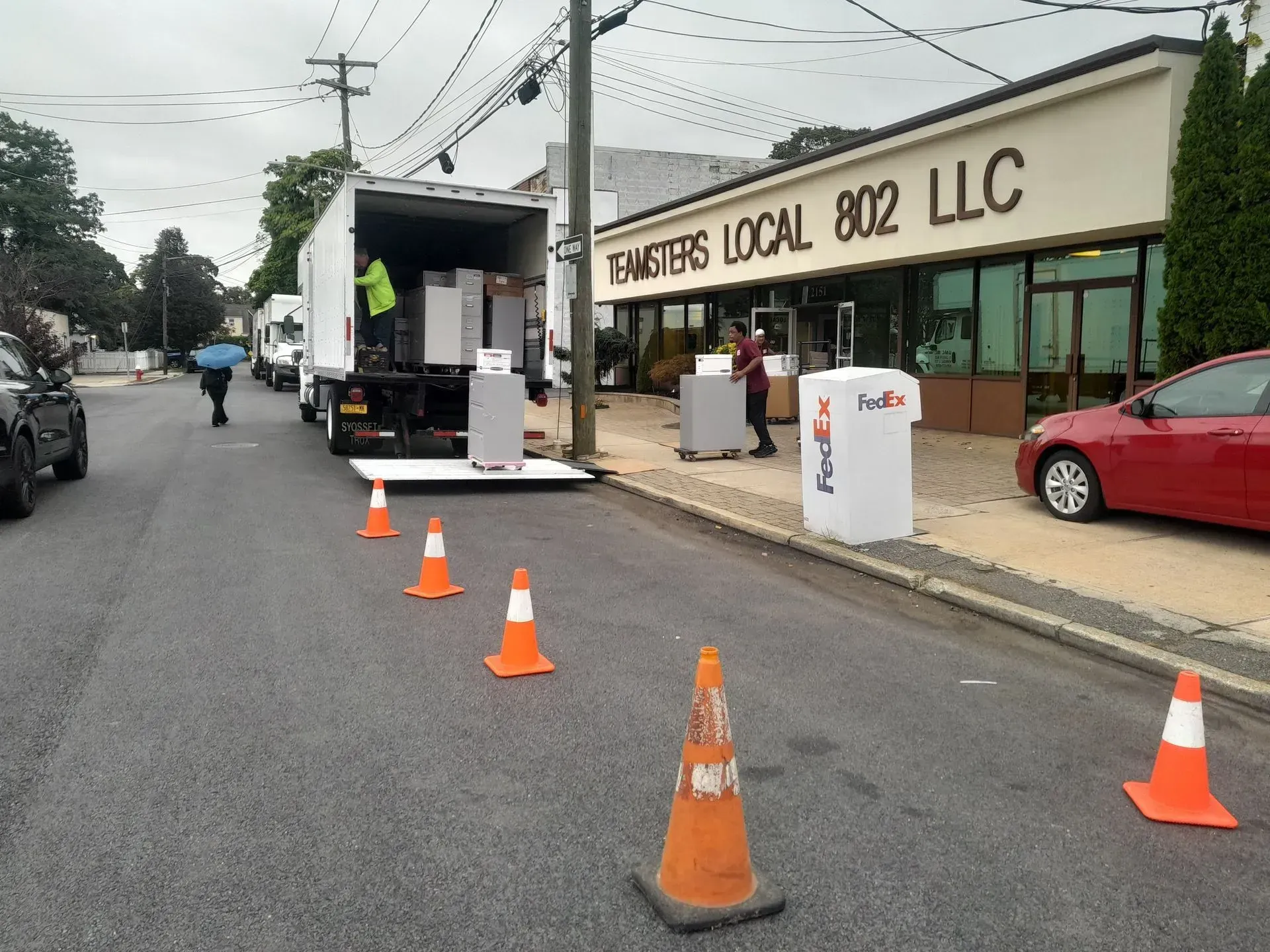 Small storefront with orange cones outside, a truck unloading, and a red car parked along the street.