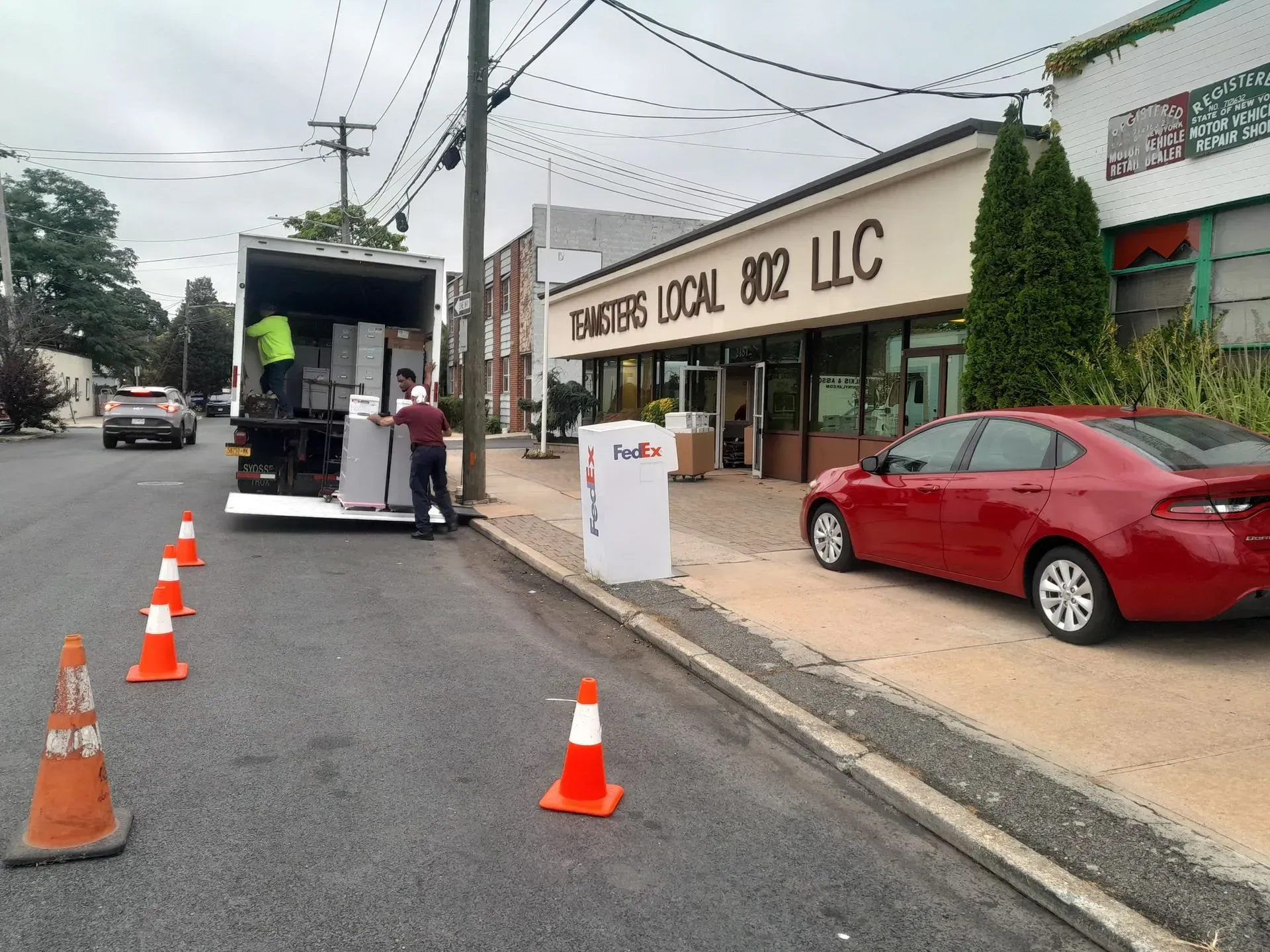 Traffic cones and a food truck on a roadside outside a red car and a storefront labeled “Food Label 302 LLC.”