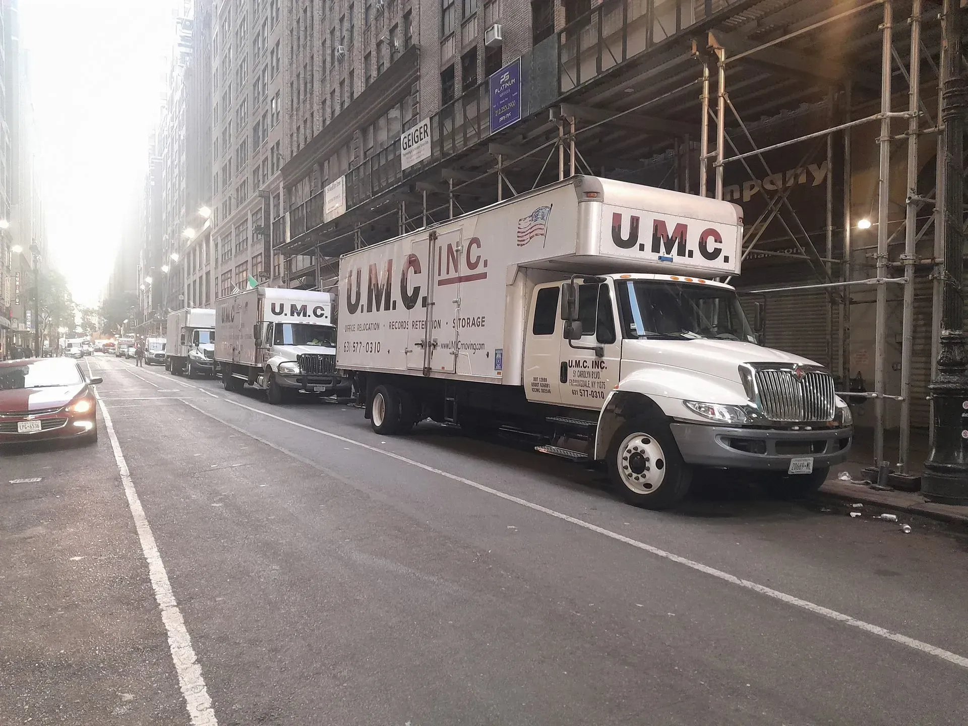 White U.M.C. moving truck parked on a city street beside scaffolding and tall buildings