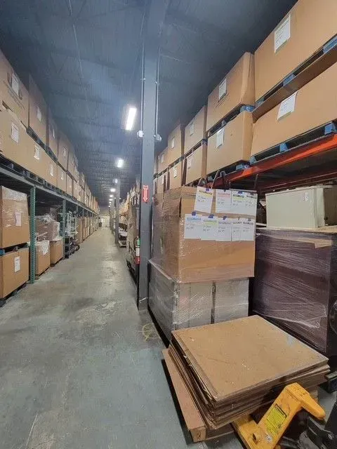 Narrow warehouse aisle lined with stacked cardboard boxes on shelves and pallets under bright overhead lights