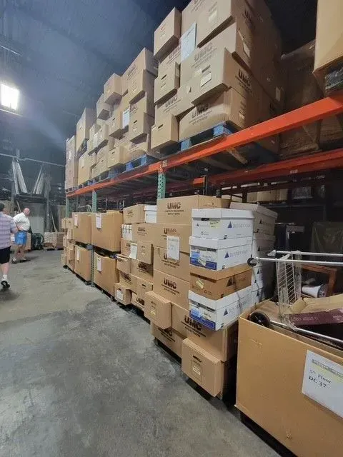 Warehouse aisle stacked with cardboard boxes on shelves and floor, with a person walking in the background.
