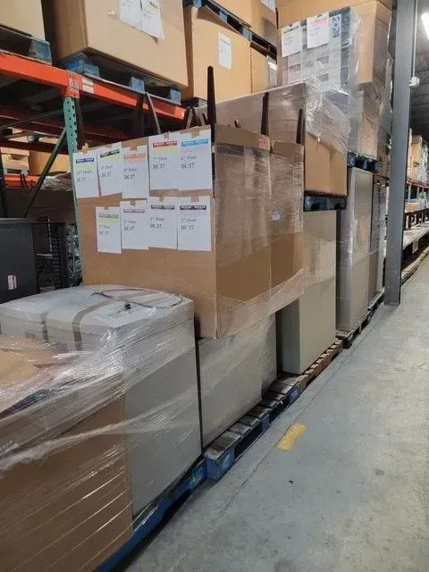 Warehouse aisle with stacked shrink-wrapped pallets, cardboard boxes, and shipping labels on metal shelving.
