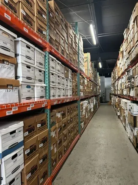 Narrow warehouse aisle lined with stacked boxes on tall metal shelves under fluorescent lights