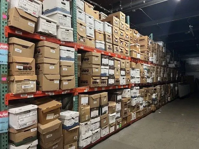Rows of stacked cardboard boxes on warehouse shelves, with a dark aisle on the right.