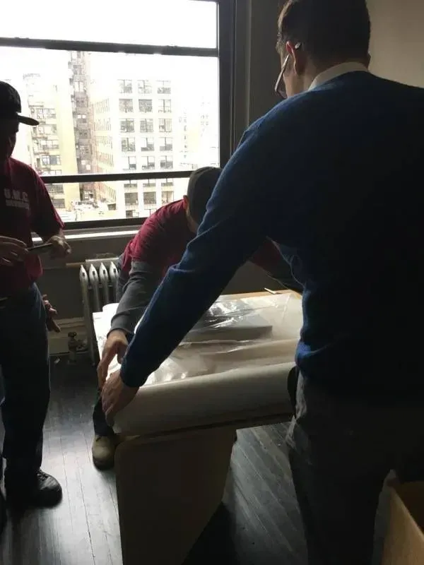 People moving a wrapped couch through a bright apartment with a large window.