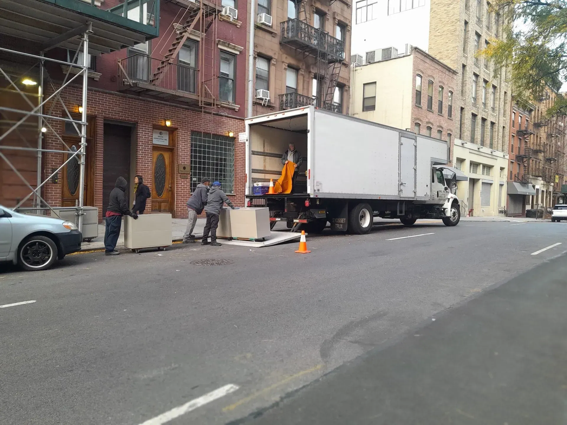Moving truck unloading on a city street beside a brick apartment building, with workers and traffic cones.