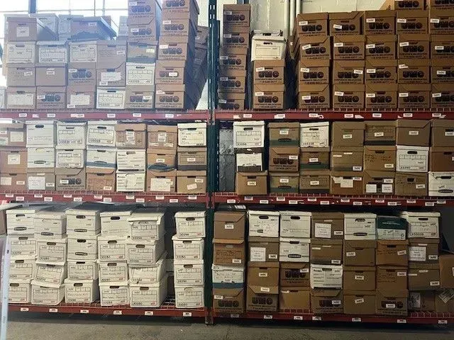 Stacks of labeled cardboard boxes on warehouse pallets, arranged in rows on metal shelving.