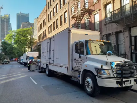 White box truck parked on a city street beside brick buildings, with a cyclist and cars in the background