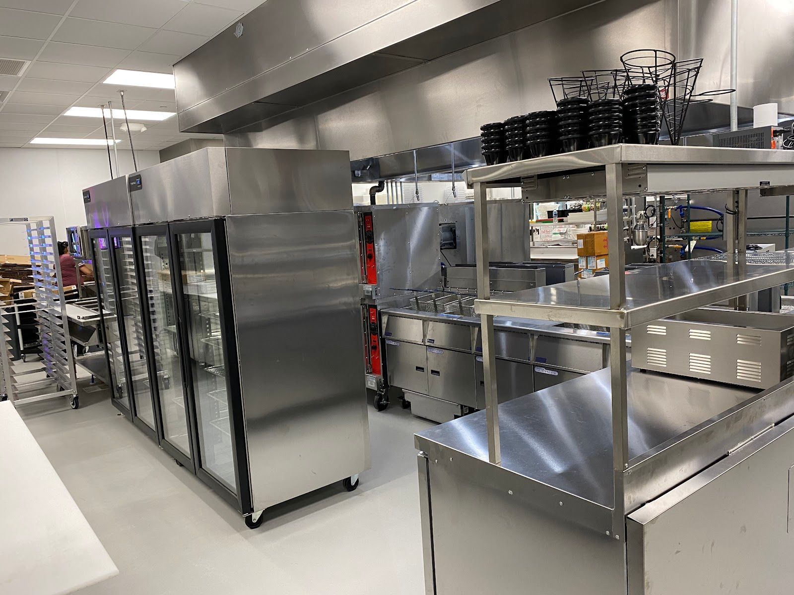 Stainless steel commercial kitchen with shelves, refrigerators, and prep counters under bright lights