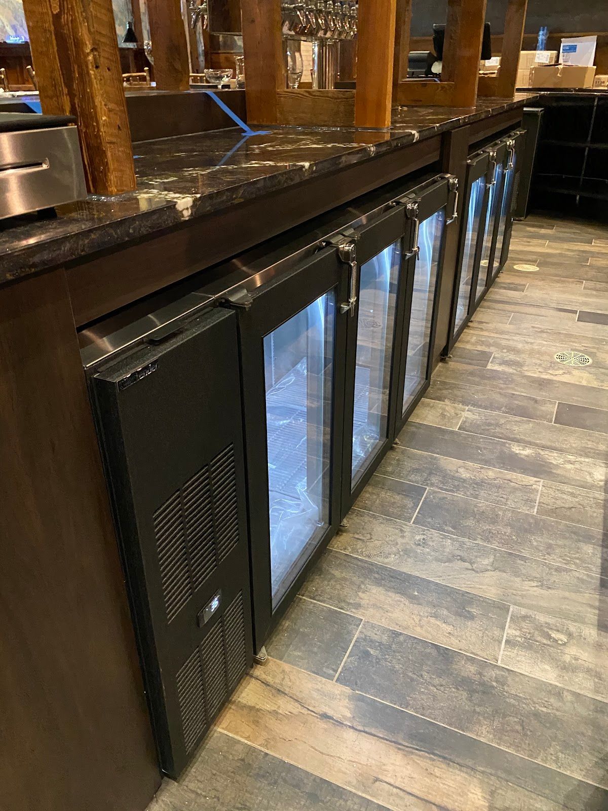 Row of black under-counter refrigerators with glass doors along a wooden bar counter.