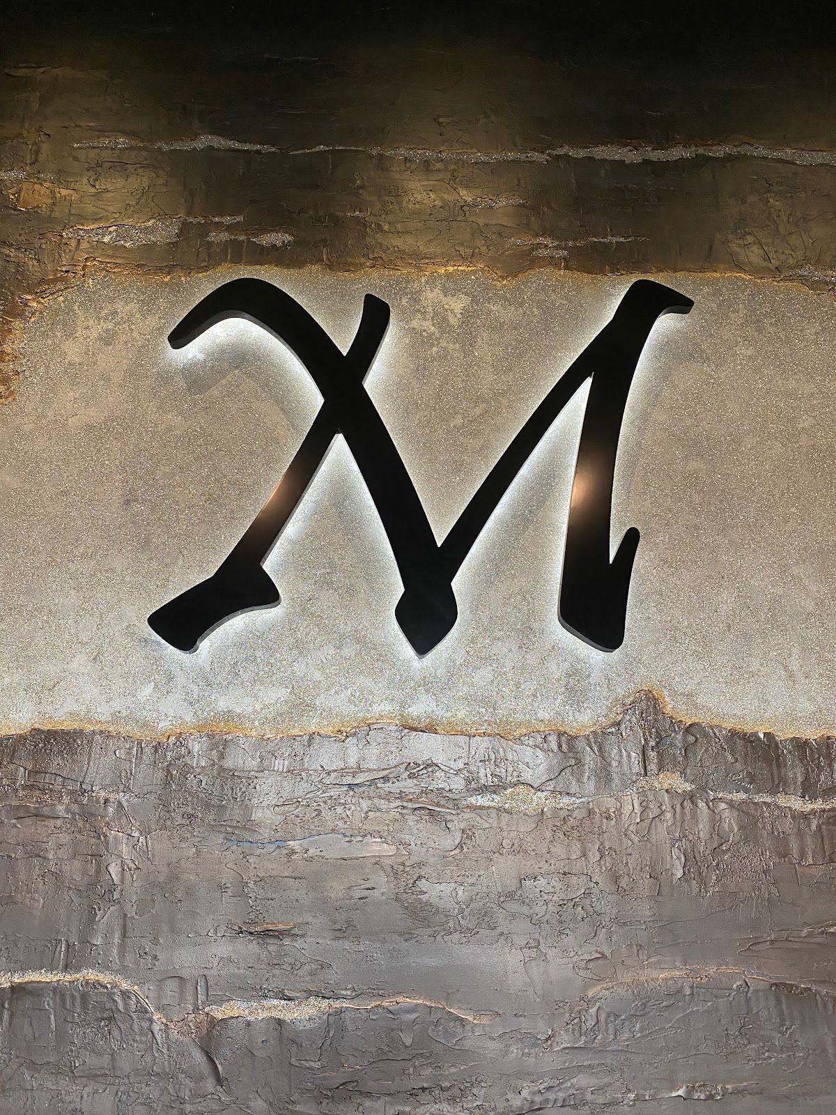 Black stylized letter M painted on a weathered gray-brown wall