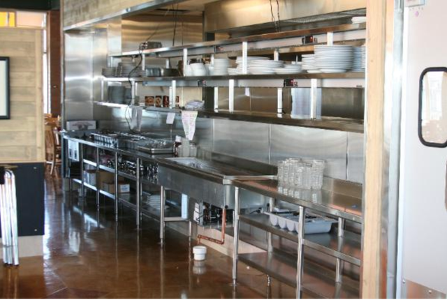 Empty commercial kitchen with stainless steel counters, shelves, and sinks against a tiled wall.
