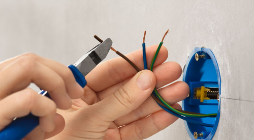 Hands strip wire with pliers next to a blue wall electrical box with exposed cables.