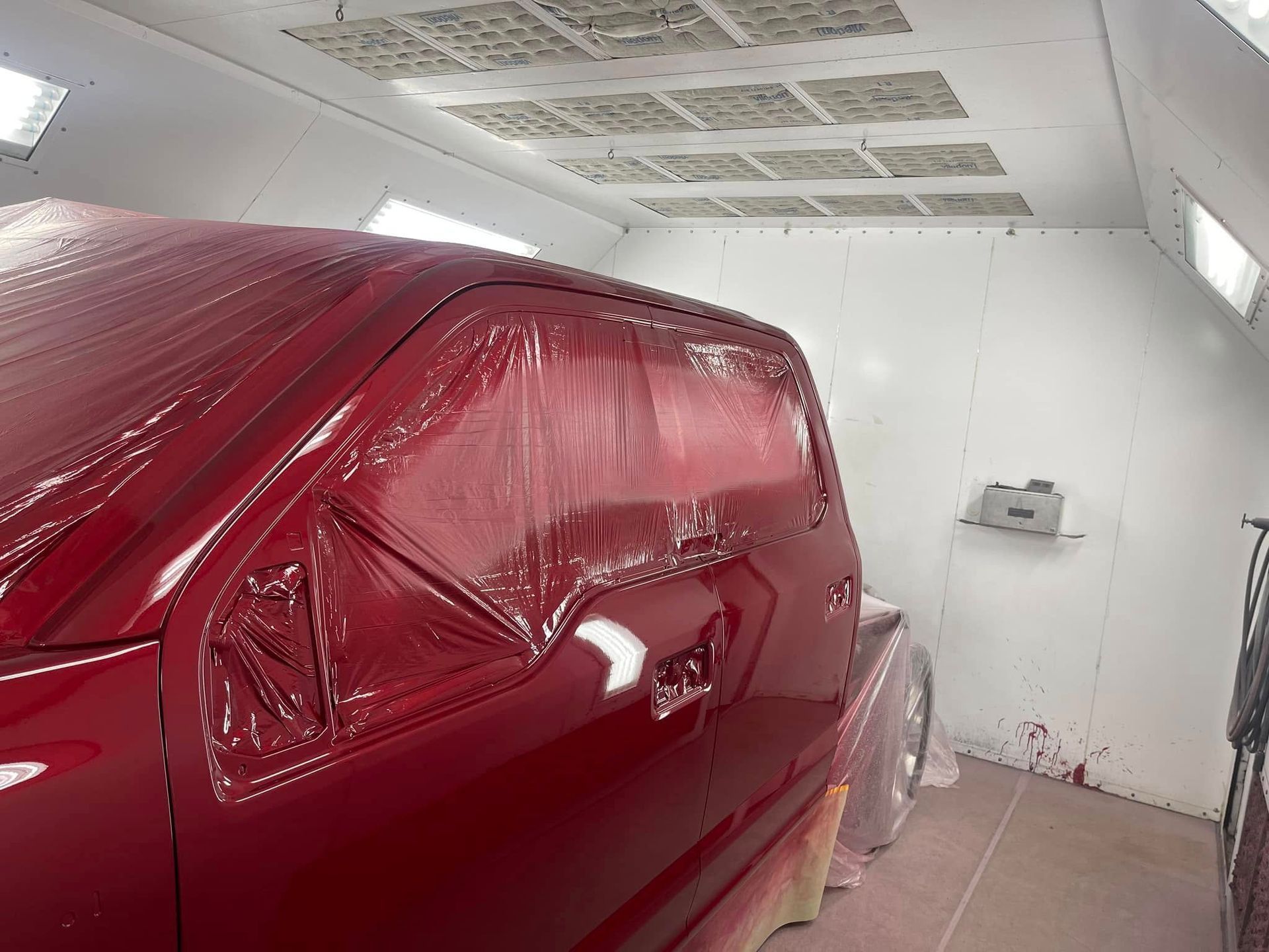 Red car body covered in plastic in a white auto paint booth