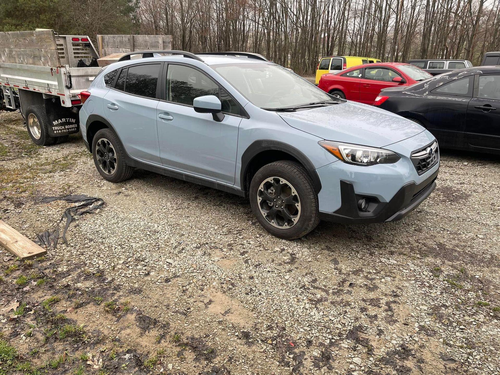 Light blue SUV parked on a gravel lot beside a flatbed truck, with bare trees in the background