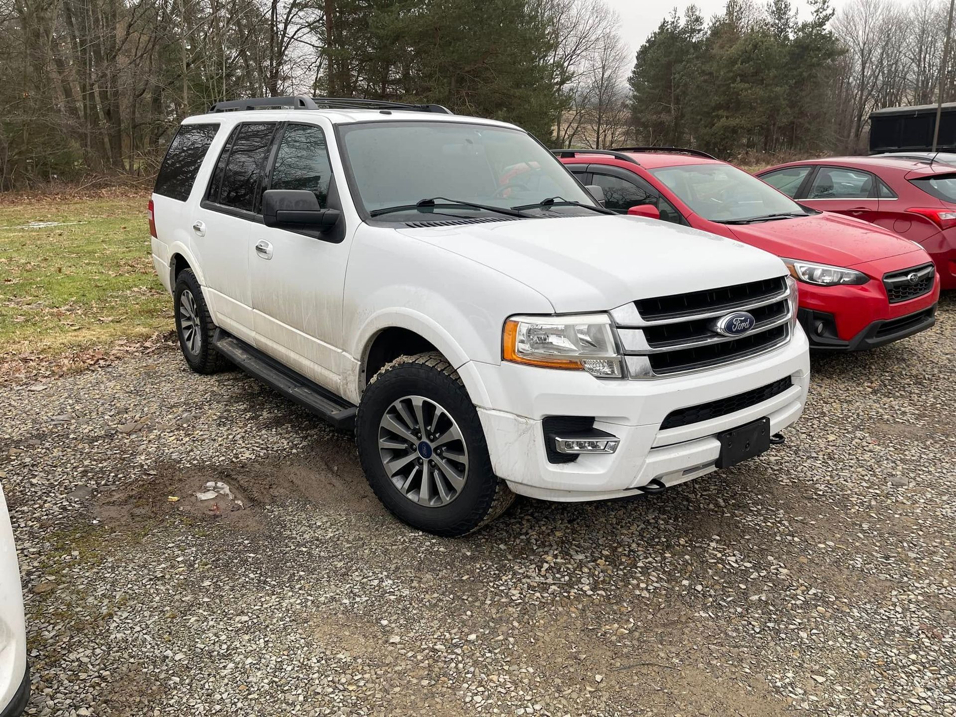 White SUV parked on a gravel lot beside a red car, with trees in the background.