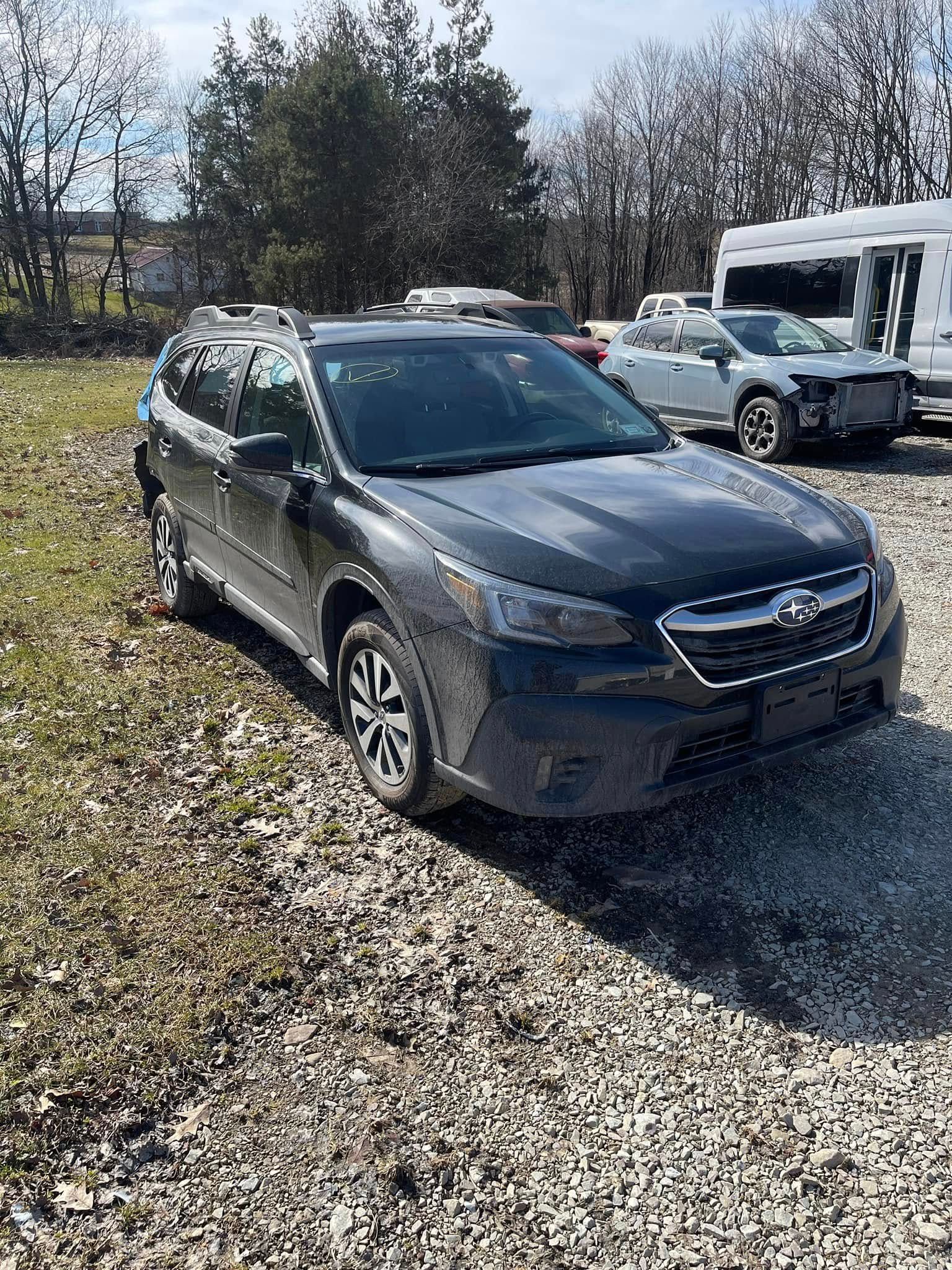 Dark gray Subaru Outback parked on gravel in a sunny lot, with trees and other vehicles in the background