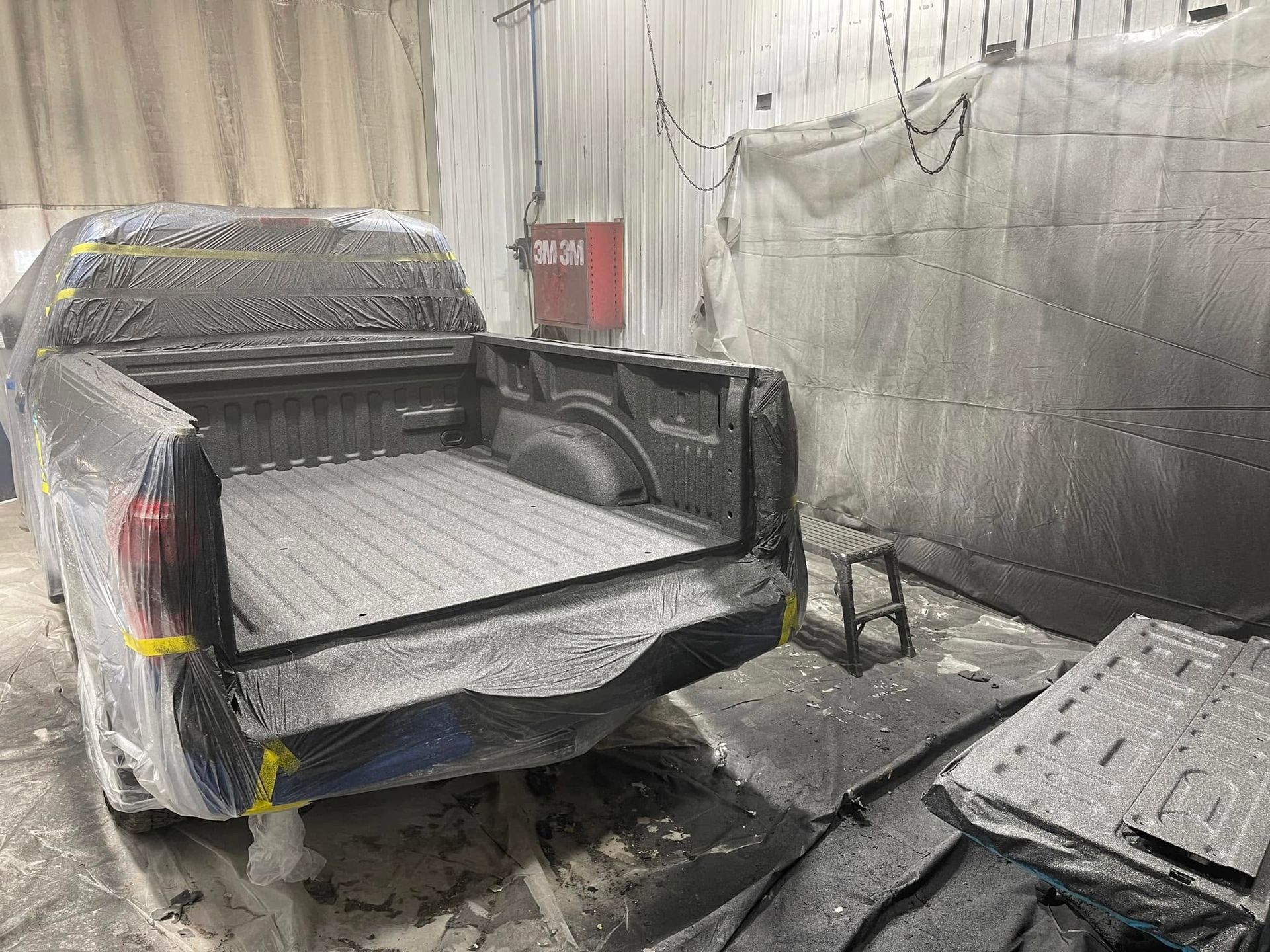 Damaged pickup truck in a paint booth with a torn bed cover and parts laid out nearby