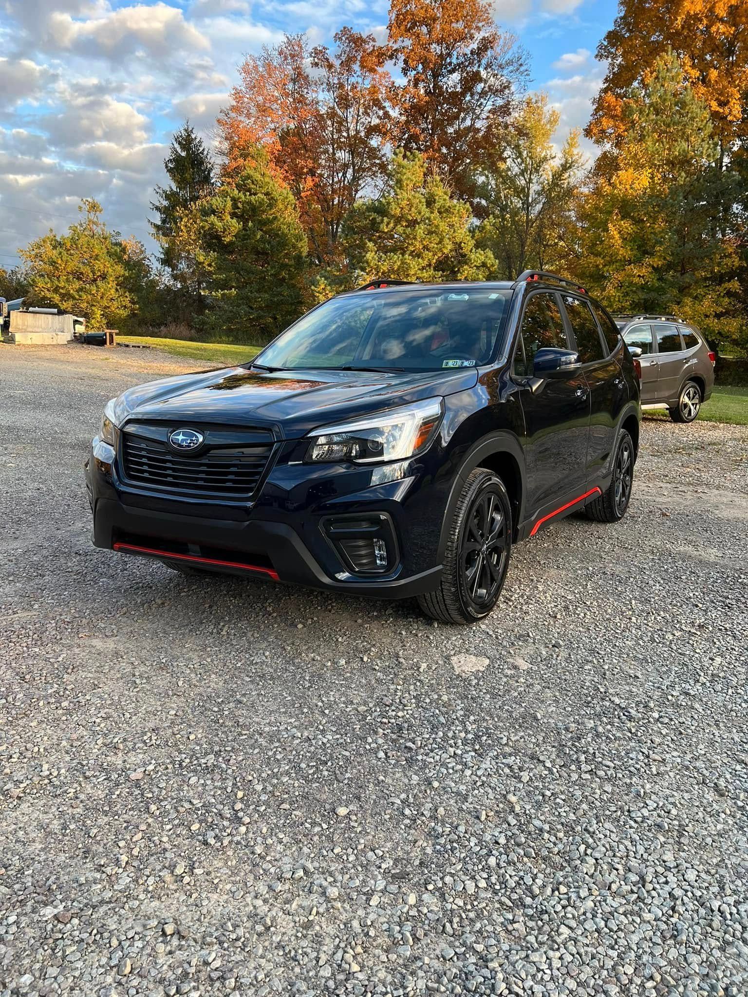 Black Subaru SUV on a gravel lot with autumn trees and a cloudy sky in the background