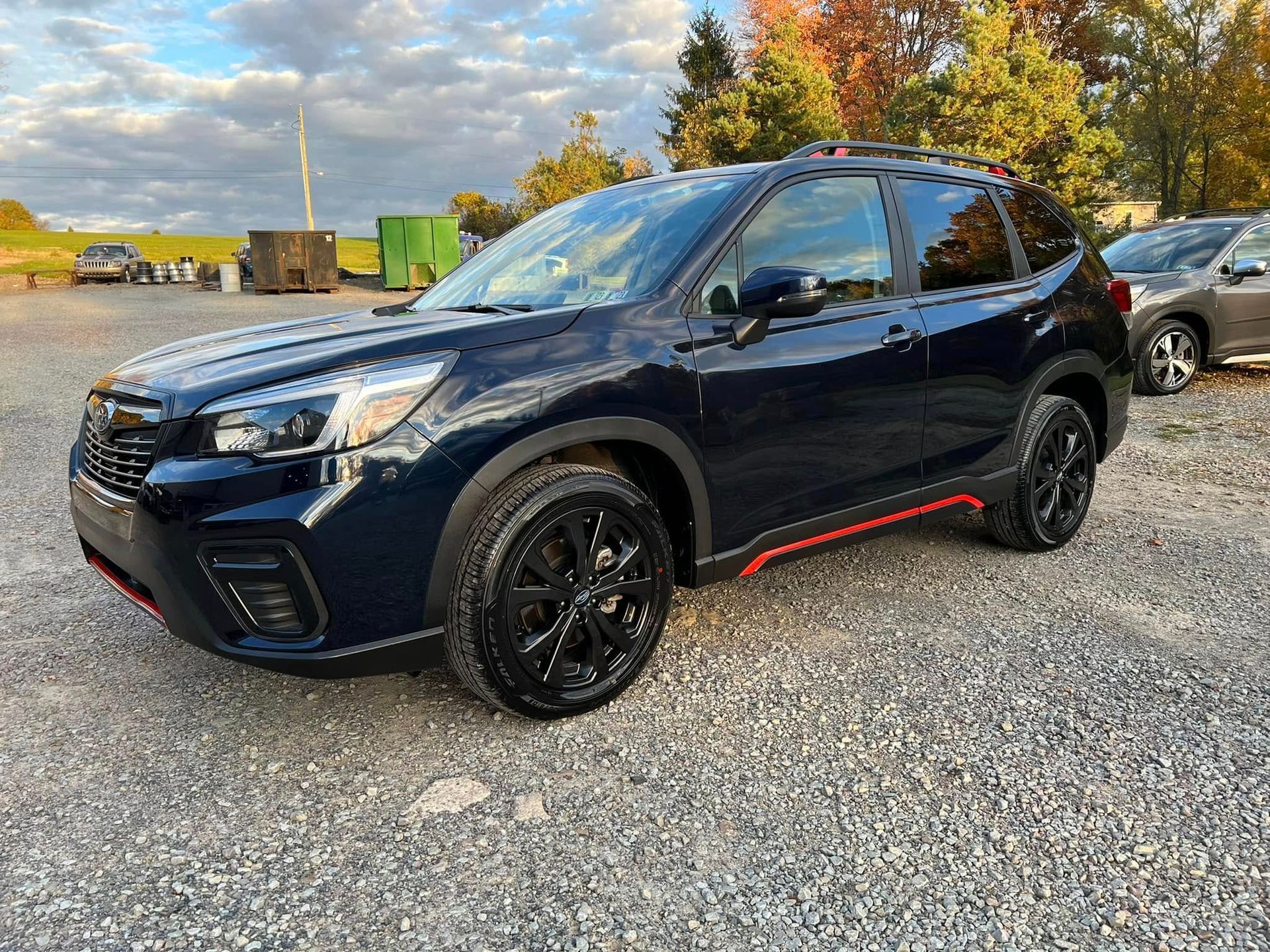 Black SUV with red side trim parked on gravel near trees and trucks under a blue sky