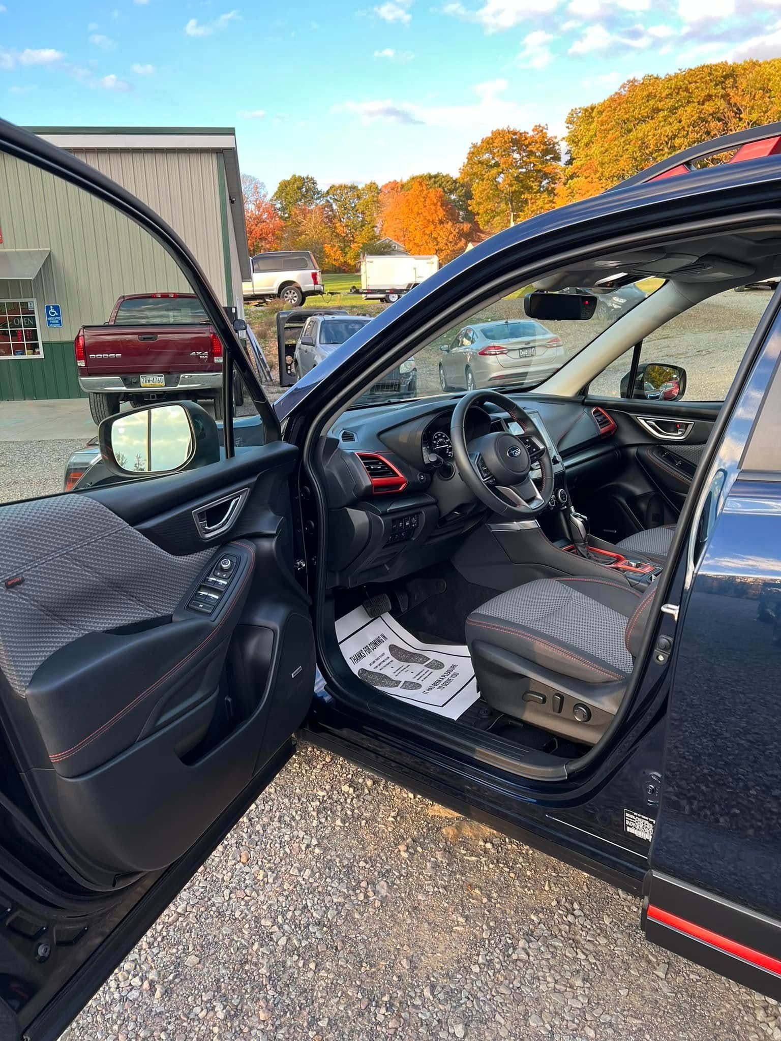 Open car door showing black interior with steering wheel and dashboard, parked outdoors under autumn trees